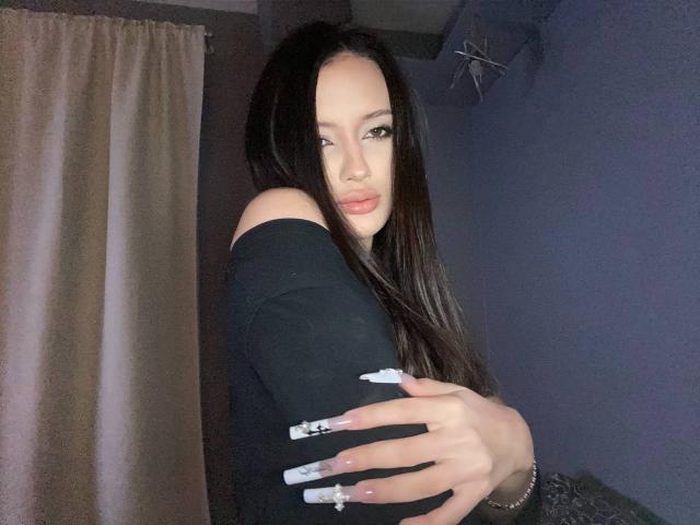 Marchelle Berrien, uma mulher atraente com longos cabelos pretos, olha sedutoramente para a câmera. Ela está vestindo uma blusa preta com um ombro de fora, com unhas longas e brancas, em um quarto com pouca luz. Muito intenso..