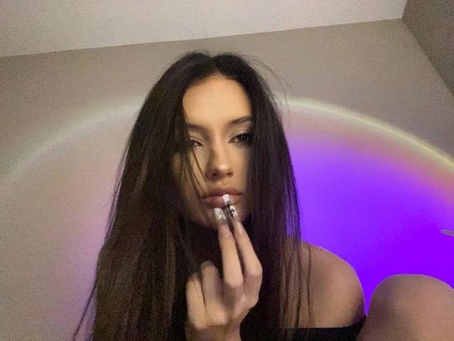 Photograph of MarchelleBerrien, dark long hair, intense gaze, holding a silver ring near her lips, purple and blue light ring in the background, sexy and provocative, shadowed face.
