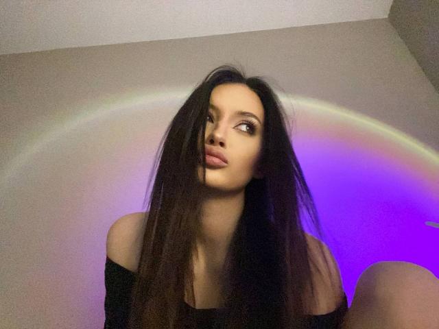 Fotografia de Marchelle Berrien, sentada com cabelos longos e pretos, usando uma blusa preta com um ombro descoberto, olhando para cima com um olhar sedutor. expressão. Arco-íris e luz azul ao fundo. Tensão sexual, pose sugestiva..