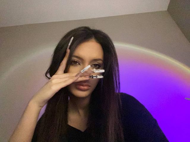 MarchelleBerrien, dark-haired woman with long straight hair, holding a small mirror in front of her face, rainbow light ring in the background, purple and blue lighting, black shirt, serious expression.