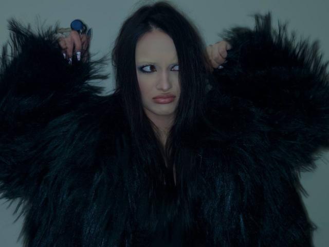 MarchelleBerrien wears a black, furry coat with arms raised, showing off its thick texture. Her blue eyes gaze sideways with a slight smirk. The lighting highlights her pale skin.