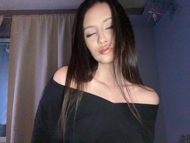 MarchelleBerrien has long, straight black hair, wearing an off-shoulder black top, eyes closed, lips slightly parted. She's standing in a dimly lit room with beige curtains in the background. Fucking sexy.