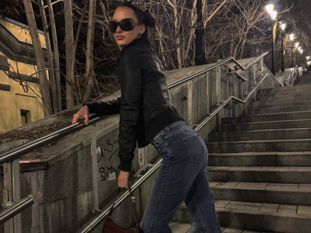 MarchelleBerrien leans against a metal railing on a dark, nighttime staircase. Wearing black sunglasses, a leather jacket, and tight blue jeans, showcasing her round ass. Streetlights and trees in background.