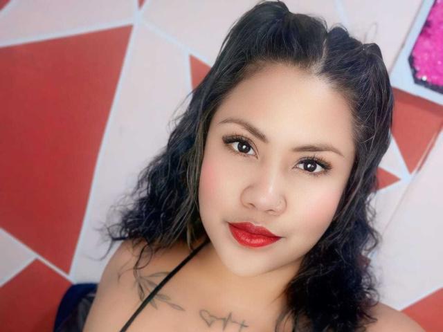 Nahaufnahme von CataleyaNoir, einer asiatischen Frau mit schwarzen, welligen Haaren, rotem Lippenstift und schwarzem Eyeliner. Sie hat Tattoos auf ihrer Brust und trägt ein schwarzes Top mit Halstuch. Im Hintergrund sind rote und weiße geometrische Muster zu sehen. Ihr Ausdruck ist selbstbewusst und leicht verführerisch..