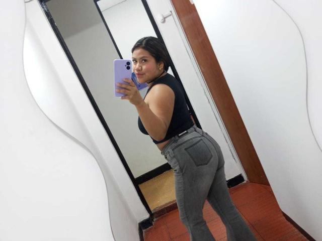 CataleyaNoir è in una stanza e sta scattando un selfie allo specchio con uno smartphone. Indossa una canotta nera senza maniche e dei pantaloni grigi aderenti, mettendo in mostra il suo sedere rotondo. Lei sta guardando indietro con un leggero sorriso..