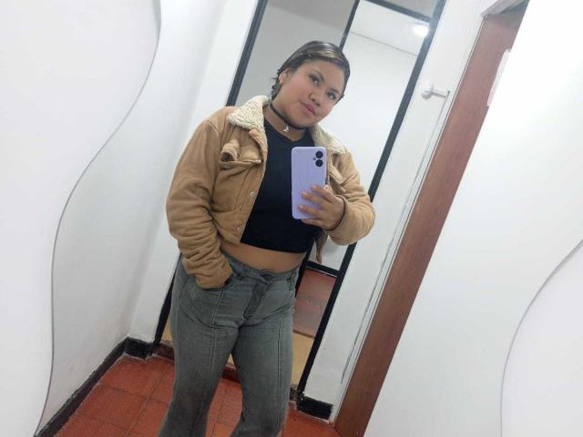CataleyaNoir takes a selfie, standing in a hallway with white walls and red carpet. She wears a tan leather jacket, black top, and blue jeans, holding a purple phone. Fucking hot.