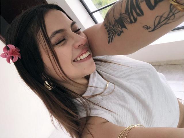 Nahaufnahme von NyraVanel mit dunkelbraunen Haaren, einem rosaen Blumen in den Haaren, lächelnd, mit geschlossenen Augen, einem weißen Top ohne Ärmel und Tätowierungen. Arm, goldene Armband, natürliches Licht, lässige Pose, entspannt, leicht provokativ..