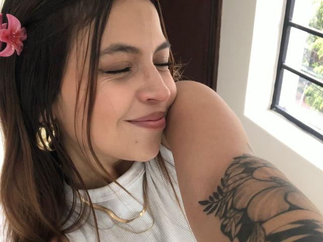 Close-up photo of NyraVanel with long brown hair, pink flower clip, and gold hoop earrings, licking the tattooed bicep of a light-skinned person, sunlight streaming through window. Fucking sexy.