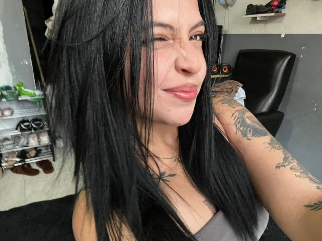 Close-up van NyraVanel, een vrouw met lang zwart haar, die knipoogt en glimlacht met rode lippen. Ze heeft tatoeages op haar arm, draagt een grijze top en bevindt zich in een kapsalon. Heel aantrekkelijk.