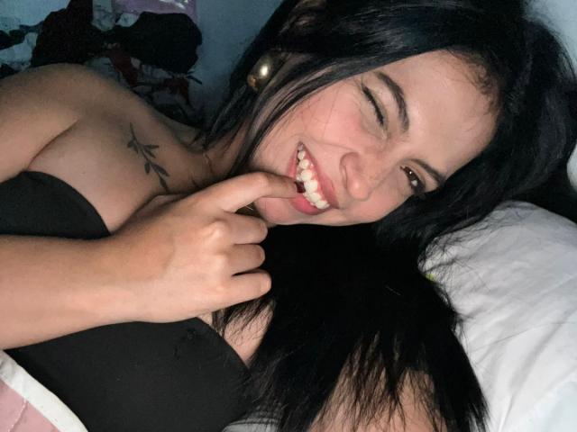 NyraVanel, com cabelo preto, sorrindo mostrando os dentes, mordendo o dedo. Ela está vestindo uma blusa preta sem alças, que revela uma tatuagem de estrela em seu ombro direito. Seus olhos estão semicerrados, com um olhar sedutor..
