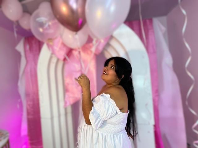 Cassandra Valmont, met lang zwart haar, lachend, gekleed in een witte jurk met open schouders, omringd door roze en witte bloemen. ballonnen, staand voor roze en witte gordijnen, doordrenkt van seksuele spanning..