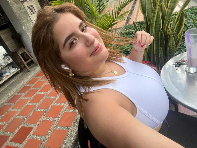 Photograph of IvyOconner, a curvy woman with light skin and brown hair, wearing a white tank top, smiling at the camera. She's sitting outdoors near a table with a glass of water. Her hand rests on her thigh, creating a subtle, sexy pose.