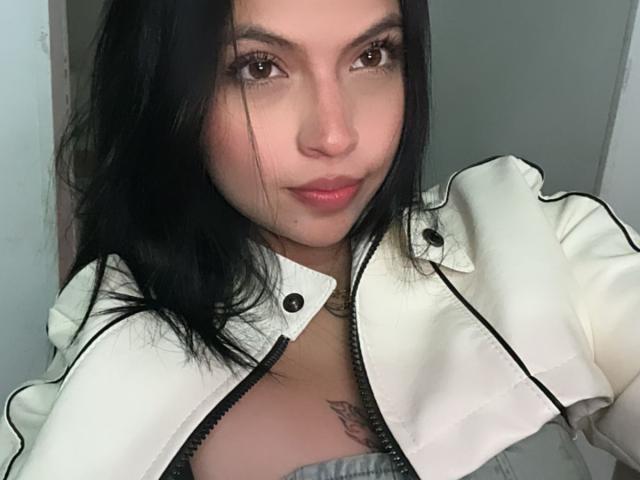 Primer plano de NyraVanel con cabello oscuro, piel clara y ojos marrones, vistiendo una chaqueta blanca abierta con detalles negros. mostrando su escote y un tatuaje en su pecho. Tensión sexual evidente..