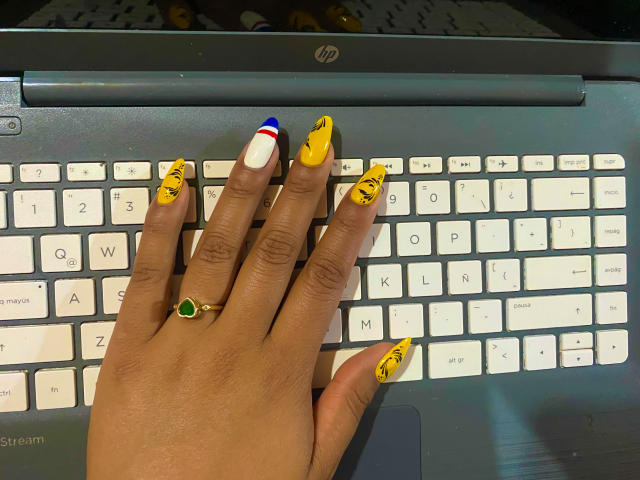 Uma mão com unhas longas e coloridas (amarelas com padrões pretos, brancas com um ponto vermelho, azuis) está apoiada em um MacBook preto. teclado. SophieSonyas usa um anel verde. Unhas incrivelmente vibrantes..