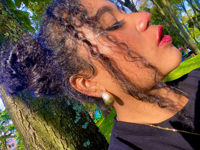 Close-up of SophieSonyas with curly black hair, red lipstick, and gold earrings. She's looking to the side, sunlight shining through leaves, casting shadows on her face. Fucking sexy, vibrant colors.