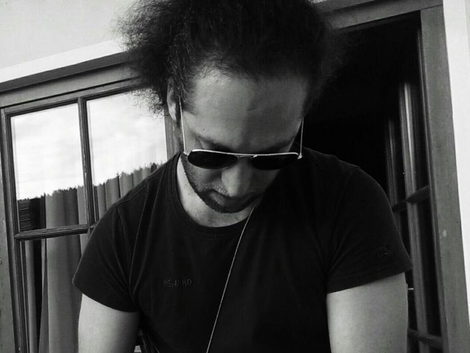Black-and-white photo of SidesHowCan with messy hair, looking down, wearing sunglasses and a black t-shirt. Slight shadow on face, hands not visible. Window with grid pattern in background.