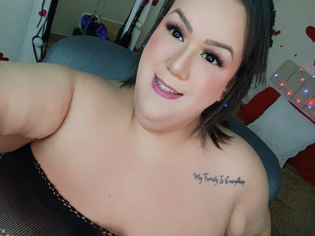Close-up van GiiannaMorrys met kort zwart haar, lichte huid en een tatoeage op haar rechter schouder met de tekst "Je hebt niet... Om groot te zijn, wees gewoon bereid. Ze glimlacht, is bloot bovenin, en haar decolleté is zichtbaar..