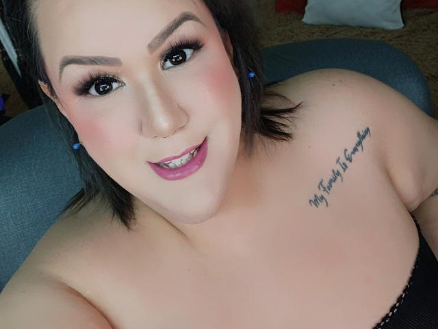 Close-up de GiiannaMorrys, uma mulher curvilínea com pele clara, cabelo preto e maquiagem marcante, sorrindo para a câmera. Ela tem uma tatuagem no ombro esquerdo e veste uma blusa preta sem alças..