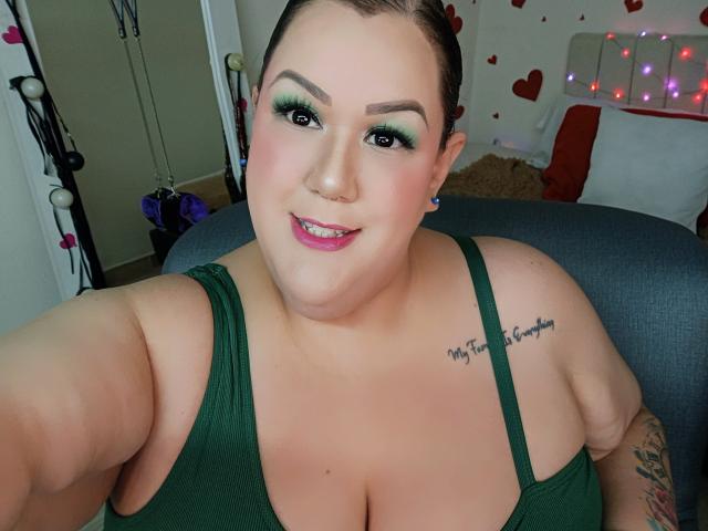 Photograph of a curvy woman, GiiannaMorrys, with dark green tank top, large breasts, tattoo on shoulder, green eye makeup, pink lipstick, and a confident smile. Dimly lit room with heart decorations.
