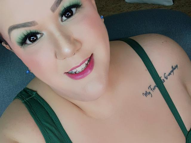 Close-up photo of GiiannaMorrys, smiling with green eyeshadow, black eyeliner, and pink lipstick. She has a tattoo on her right shoulder reading "My heart is the sunshine". Wearing a green strap top.