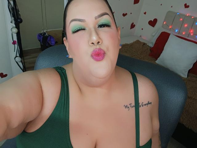 Photograph of a chubby woman with green eyeshadow, pink lipstick, and a nose ring, blowing a kiss. She wears a green tank top, exposing her cleavage and "My Forever You" tattoo.