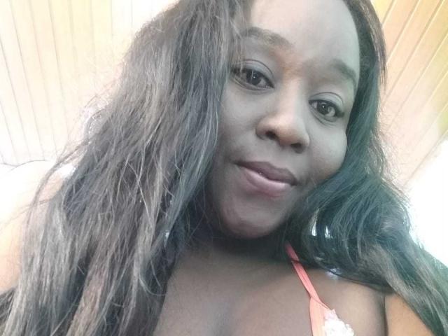 Selfie em close-up de Yirily, uma mulher negra com cabelos longos e lisos castanhos, vestindo um top vermelho de alças finas, com os lábios levemente curvados para dentro. e um sorriso suave, luz brilhante vinda de cima, leve sombra no rosto..