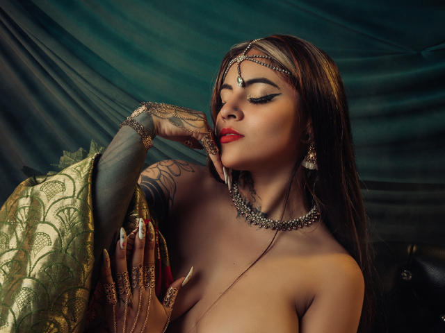 CoriPaige with closed eyes, dark brown hair, red lipstick, wearing gold jewelry and sheer black glove, topless, with gold embroidered cloth draped over shoulder, dark background, sexy, seductive pose.