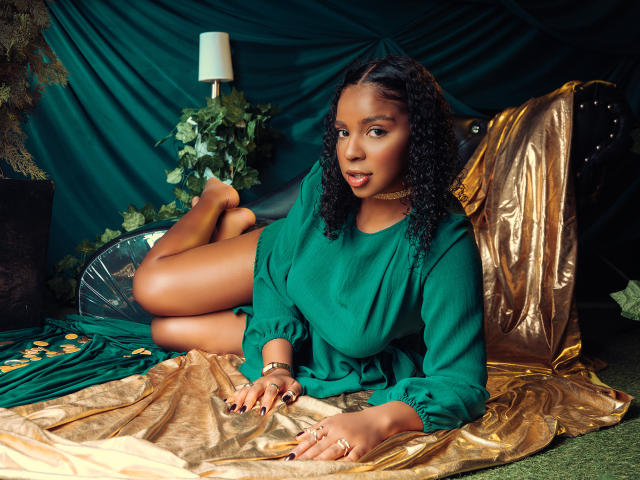 LisaMendez lies on a gold satin sheet, legs bent and raised, wearing a green blouse. Her dark curly hair contrasts with the green and gold backdrop. Sexual tension is palpable.