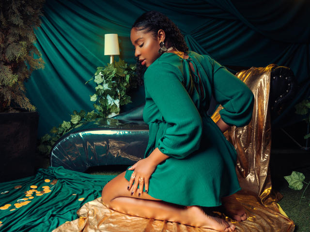 LisaMendez, donkerhuidig, met krullend haar, knielend op een gouden folie, gekleed in een groene jurk, zijaanzicht, verleidelijk. blik, hand op de dij, zacht licht van een lamp, donker blauw-groene achtergrond..