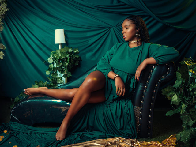 LisaMendez, à la peau foncée, aux formes généreuses, vêtue d'une robe verte, est assise en retrait sur une chaise noire ornée de clous, une jambe croisée par-dessus l'autre. autre.
elle regarde vers le bas, avec une lampe et des plantes en arrière-plan..