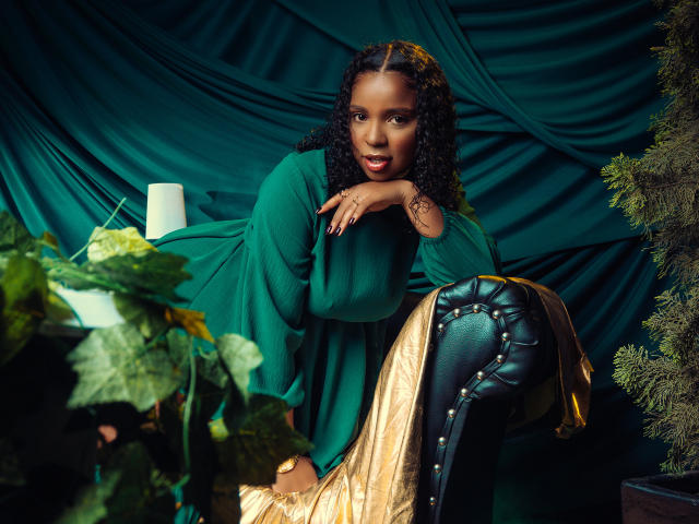 LisaMendez, dark-skinned with curly hair, leans on a black leather chair, wearing a green robe, looking seductive. Plants and green drapery in the background. Dim lighting, intense gaze.