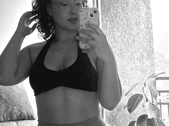 Black-and-white selfie of SophiaBrowm, curly hair, glasses, holding phone with dual camera, wearing black sports bra, hand in hair, potted plant in background, strong light source, confident pose.