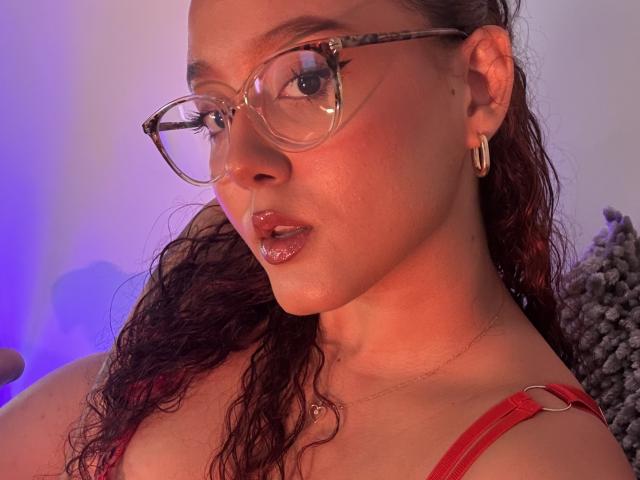 Close-up of SophiaBrowm, a sexy woman with curly brown hair, wearing red strap top and clear glasses, looking at camera with red lighting. She's got hoop earrings and a slight smirk.