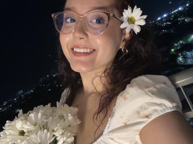 Close-up of SophiaBrowm, a smiling woman with curly dark hair, wearing glasses and a white dress with puffed sleeves. She holds white daisies, one in her hair. Nighttime city lights in background.