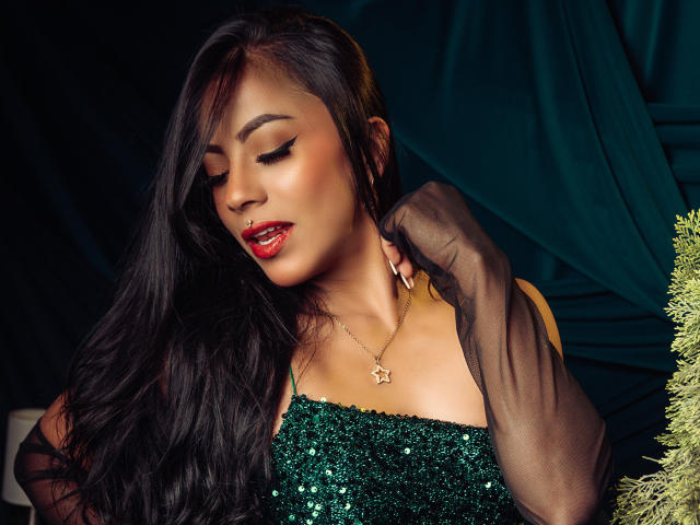 Foto van Amberv, een Aziatische vrouw met lang zwart haar, die een groene jurk met pailletten en transparante zwarte handschoenen draagt. Haar schouder vasthoudend. Ze heeft rode lippenstift en een gouden ketting. Verdomd sexy..