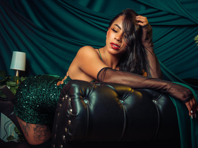 Amberv, com a pele escura e o cabelo preto comprido, inclina-se de forma sedutora em um sofá de couro preto. Vestindo um vestido verde brilhante e luvas pretas transparentes, transmitindo uma intensa tensão sexual. Lâmpada ao fundo.