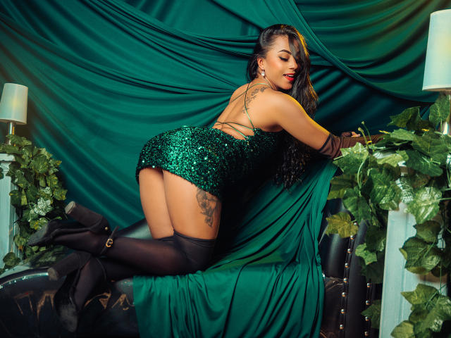 Amberv, uma mulher curvilínea com pele escura e batom vermelho, ajoelha-se em um sofá de couro preto, vestindo uma saia verde com lantejoulas. Botas pretas que chegam até a coxa, e luvas que chegam até o cotovelo. Cortinas verdes e plantas ao fundo.