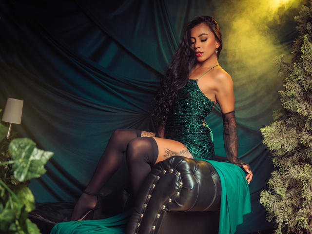 Amberv è seduta su una sedia, indossa un abito verde scintillante, stivali neri alti fino alla coscia con borchie e lunghi guanti. È attraente, con i capelli scuri intrecciati e un'espressione sensuale. Una luce gialla brilla dietro di lei
.