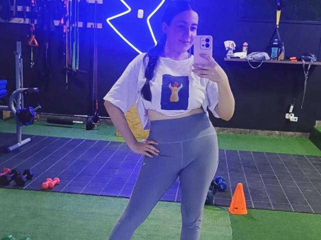 MabyLegrand tira uma selfie em uma academia, vestindo uma cropped branca com uma estampa de chave e leggings cinzas justas. Luzes azuis neon formam um raio atrás dela. Incrivelmente atraente..