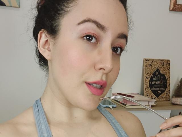 Primer plano de MabyLegrand con cabello oscuro, lápiz labial rosa y camiseta azul claro, sosteniendo un tenedor cerca de la boca, piel clara. Ligeras mejillas enrojecidas, fondo minimalista con elementos decorativos de madera. Increíblemente atractivo/a..