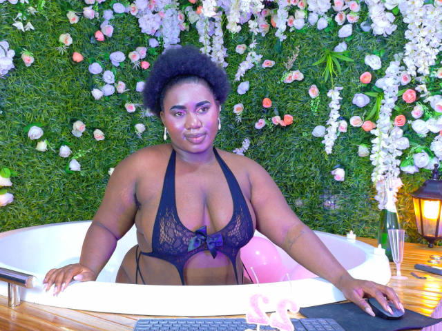 Dilay with dark skin and curly hair, wearing a black lace halter top, sitting in a white bathtub. Floral green wall background, keyboard on wooden floor, sexy and relaxed.