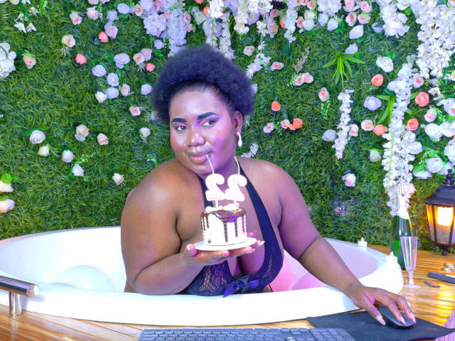 Dilay, con la pelle scura e i capelli afro naturali, è piegata su una vasca da bagno bianca, tenendo una candela accesa vicino alle labbra. Sfondo con motivi floreali. Tastiera su pavimento di legno. Tensione sessuale.