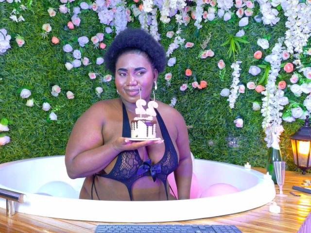 Dilay, une femme noire aux formes généreuses avec une coiffure afro volumineuse, est assise dans une baignoire blanche, vêtue d'un soutien-gorge en dentelle noire, et tient un gâteau. Contexte : mur vert avec des fleurs blanches. Suggestif, sexy..