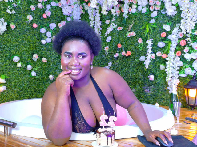 Arte digital fotorrealista de Dilay, uma mulher negra curvilínea com seios grandes, vestindo um top de renda preto com alças finas, apoiada em uma mesa branca com uma parede verde com flores ao fundo. Com um sorriso malicioso, dedo nos lábios..