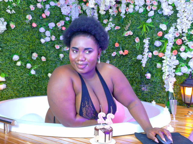 Dilay, una donna nera di taglia comoda, con una figura formosa e un seno abbondante, è sdraiata in una vasca da bagno bianca, indossando un reggiseno in pizzo. Sfondo con parete verde decorata con fiori. Uno sguardo intenso..