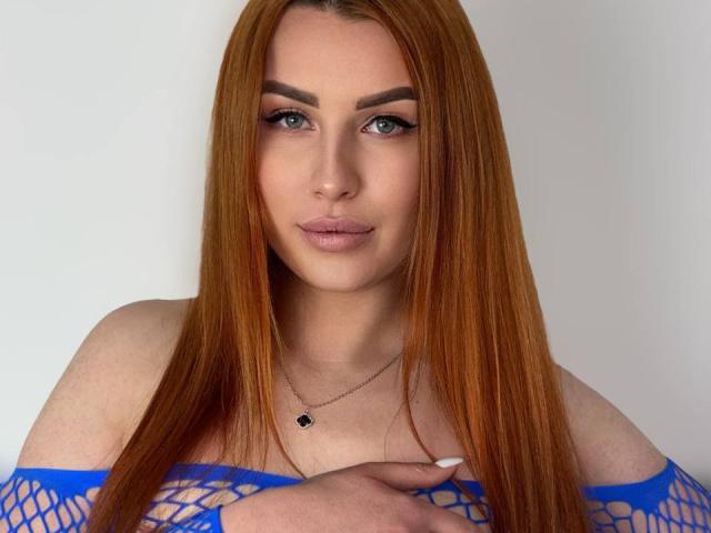 Close-up of KiraNeeww with long, straight red hair, wearing a blue fishnet top, pale skin, full lips, blue eyes, and a small black pendant necklace. Her hand covers her chest.