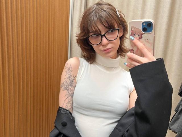 ErynAttridge, cabelo castanho curto, óculos de aro preto, blusa branca sem mangas, braço tatuado, segurando um telefone com capa rosa, jaqueta preta com um ombro descoberto, expressão levemente emburrada, iluminação suave, selfie casual..