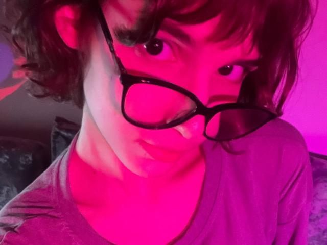 Photo en gros plan d'ErynAttridge, avec des cheveux bruns courts et des lunettes à monture noire, penchée en avant avec une expression intense. Un regard avec une légère teinte rosée.
l'éclairage rose et violet crée une atmosphère provocante et érotique. Cou et poitrine apparents.