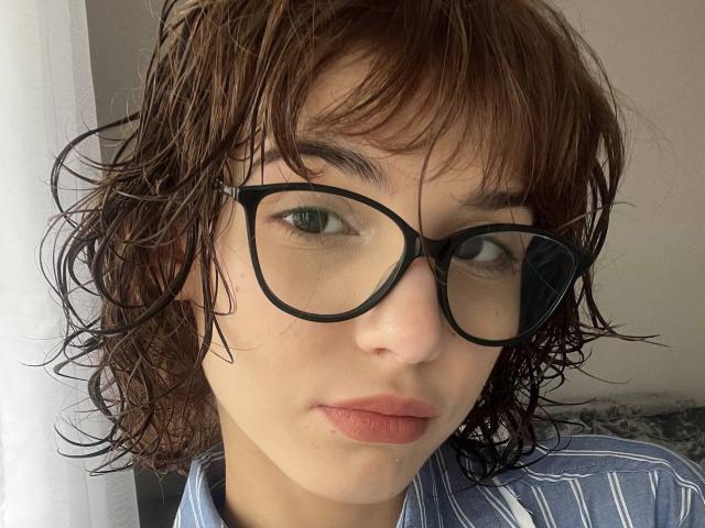 Close-up photo of ErynAttridge with wet, dark brown, shoulder-length hair, wearing black-framed glasses and a blue-striped shirt. ErynAttridge's green eyes gaze directly at the camera, lips slightly parted.