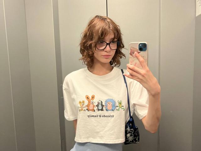 ErynAttridge, avec ses cheveux bruns hirsutes, ses lunettes et son t-shirt blanc représentant des lapins de dessin animé et le texte "Hello 3D World". se prend un autoportrait dans un miroir. Elle tient un téléphone avec une coque bleue..